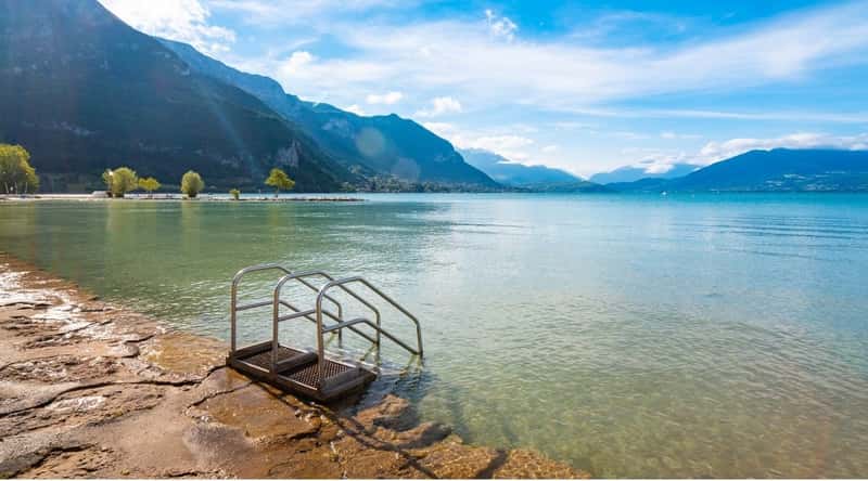 View of Annecy's beach in sunlight. Along the pristine shores of Lake Annecy, these sandy stretches are bordered by lush greenery and offer striking views of the surrounding French Alps. The water in the lake is crystal clear with shades of turquoise and emerald.