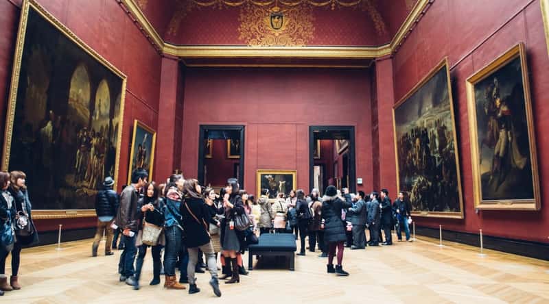 An interior view of one of the halls within the Louvre Museum in Paris, France. The hall is characterized by dark burgundy colors and adorned with large paintings on the walls. Numerous visitors are observed looking at the artwork.