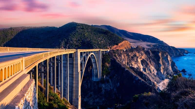 The image features Bixby Bridge, also known as Rocky Creek Bridge, along the Pacific Coast Highway near Big Sur in California, USA, captured at sunset. The bridge is an iconic concrete arch structure spanning over a rugged canyon, providing a scenic route for coastal travel.