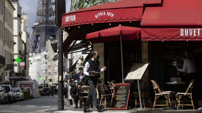 A waiter serves beverages on a cafe's terrace near the Eiffel Tower. The roof of the restaurant and the awnings on the terrace are red with white inscriptions with the name and year of opening of the restaurant. The terrace has chairs and tables for visitors, as well as a large menu board.