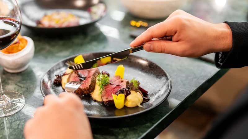 A close-up photograph depicting a dish with meat placed on a table in a restaurant setting. Hands are visible holding a knife and fork over the plate.