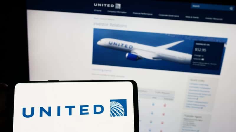 The image depicts a person holding a mobile phone with the logo of the American company United Airlines Holdings Inc. displayed on the screen. The background shows a generic web page interface.