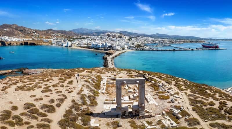 An aerial view capturing the renowned Portara Gate on Naxos island in the Cyclades, Greece. The image features the ancient stone structure set against the backdrop of the town and harbor. The Portara Gate, a prominent historical landmark, is visible with its distinctive architectural details.