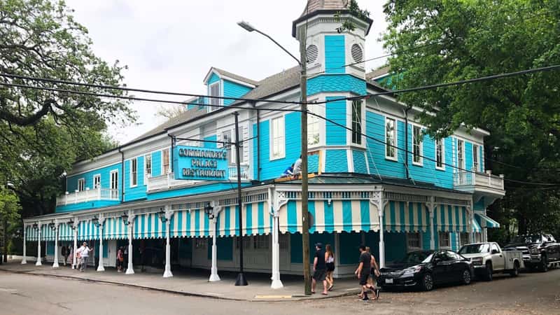 Commander's Palace in the Garden District of New Orleans features a turquoise building with white window frames. The street is lined with several parked cars, and people can be seen going about their activities. The sky appears overcast, adding a subdued atmosphere to the scene.