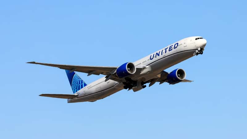 The image shows a United Airlines plane flying in the sky, featuring the airline's name "United" inscribed on its body. The plane is predominantly white with a blue tail.