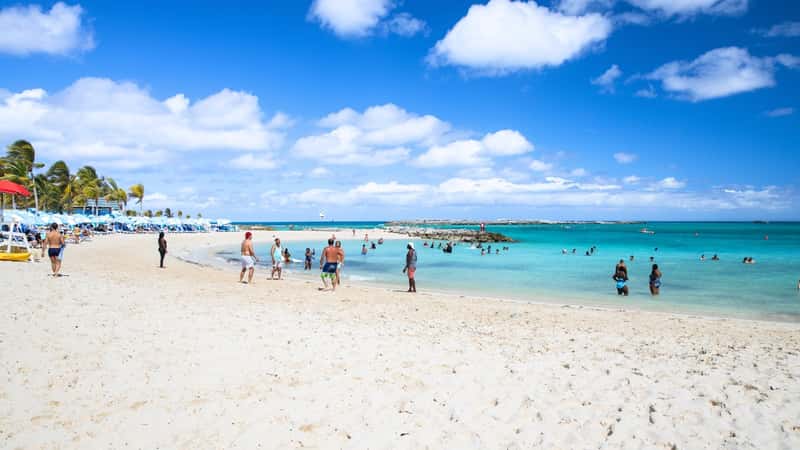 On Royal Caribbean's private island, beachgoers engage in various activities like playing ball, swimming, and sunbathing on sun loungers. The scene includes umbrellas and palm trees.