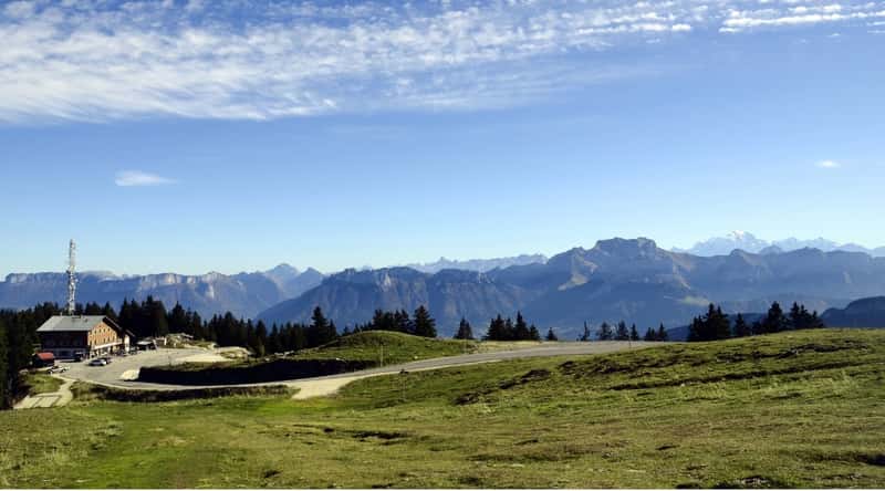 An awe-inspiring vista of Semnoz Peak, a mountain in the French Alps. The photo reveals the summit of the mountain, with a meandering road leading to a brick building, set against a backdrop of abundant pine trees. In the distance, one can also discern a multitude of mountain ranges.