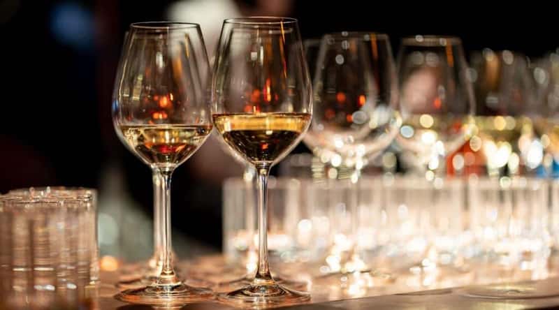 A photograph depicting two filled glasses of wine on a bar counter, surrounded by numerous empty glasses. The image conveys a scene of a bar setting with a focus on the beverages served. 