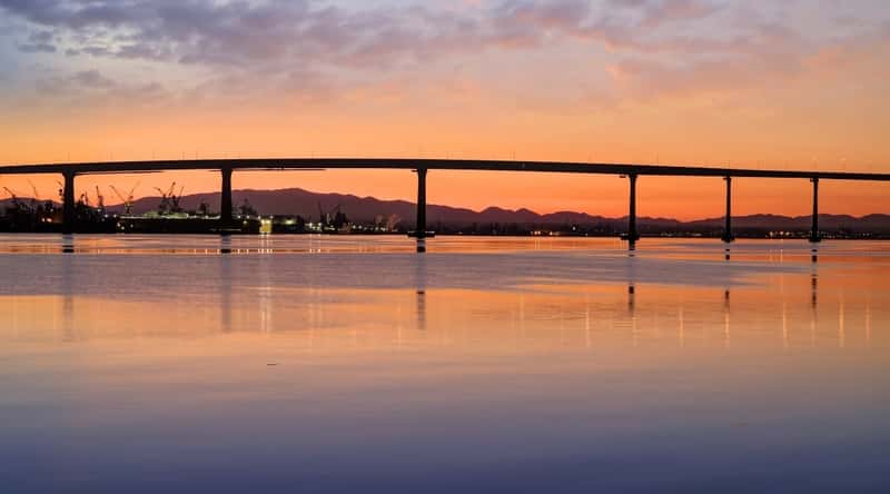 A serene photograph capturing a beautiful sunset scene overlooking the Coronado Bridge. The image features calm waters reflecting the fading sunlight, with the bridge spanning the waterway. The sky exhibits a slight cloud cover.