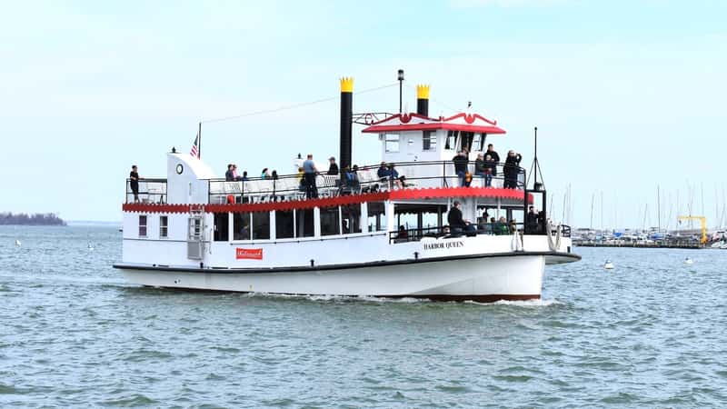 A photograph depicting a two-story white and red ship with passengers on board. The image captures the vessel's exterior, highlighting its color scheme and design. Passengers can be seen on various decks.