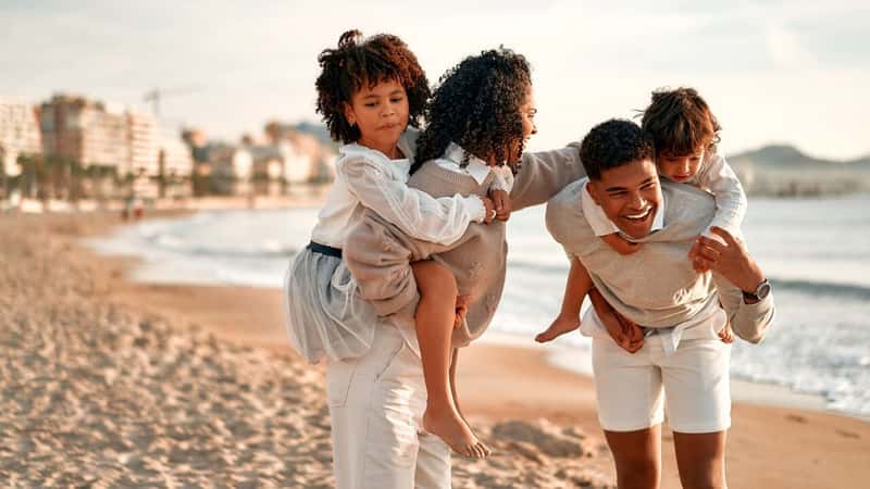 African American family with a cute curly daughter in a white dress and a little son on their backs walking and having fun on the sandy beach by the sea at dawn.