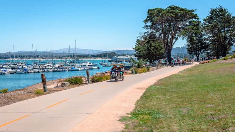 People enjoy a walk in Monterey Bay Coastal Recreation Trail on a sunny day, with the Old Fisherman's Wharf and many boats in the background.