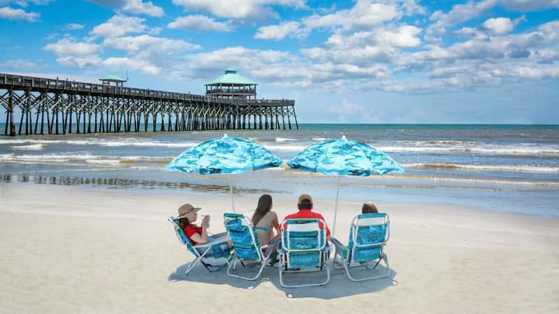 Family relaxing on the beautiful beach, People enjoying summer vacation by the ocean. Family sitting under beach umbrella. Cloudy sky and pier in the background. Folly Beach, South Carolina USA.