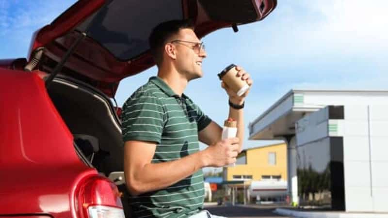 Young man with hot dog drinking coffee near car at gas station
