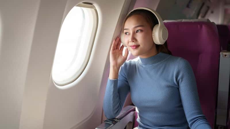Young woman wearing headphones listening to music while traveling Sitting near the window in First Class during the flight. Traveling. Connect to the WiFi Internet with your smartphone on the plane.