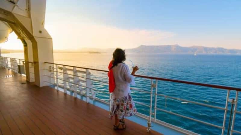 A woman sips a drink on the deck of a cruise ship as the sun sets and the ship passes islands on the Aegean Sea