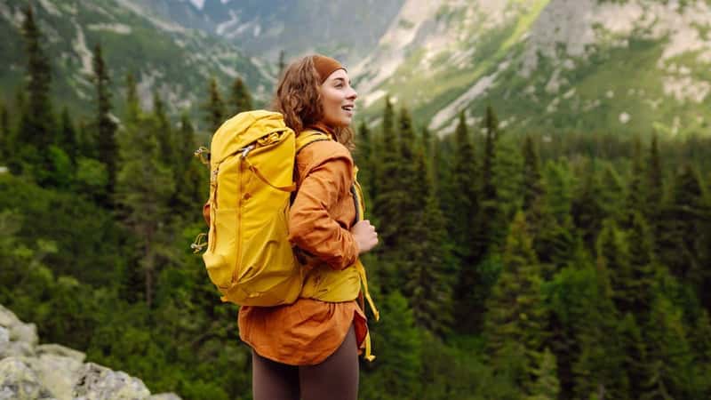 A smiling female traveler with a yellow hiking backpack enjoys the beautiful scenery of the majestic mountains. Travel, adventure. Concept of an active lifestyle.