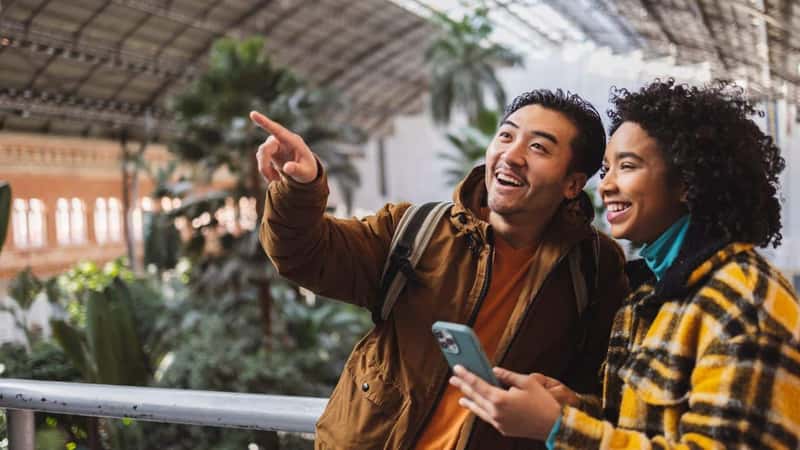 Two friends enjoying an indoor botanical garden, one pointing at a feature while the other holds a smartphone. The atmosphere highlights travel, friendship, and exploration