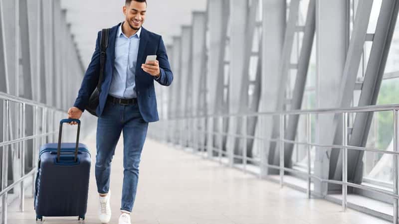 Online Check In. Handsome Arab Man Walking With Suitcase In Airport And Using Smartphone, Smiling Young Middle Eastern Guy Browsing Internet On Cellphone While Going To Flight Gate, Copy Space