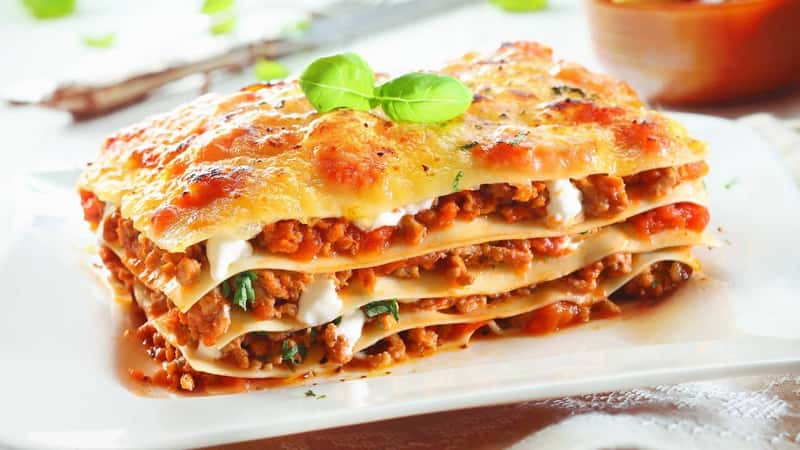 Close-up of a traditional lasagna made with minced beef bolognese sauce topped with basil leafs served on a white plate