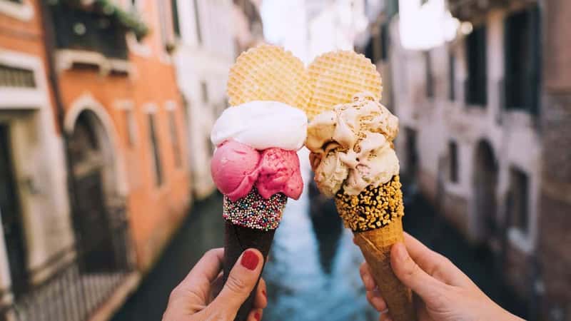 Beautiful and delicious italian gelato in waffle cone in front of streets and bridges of Venice. Ice cream in girls hands on background of water canal and historic buildings of Italy. Travel Europe.