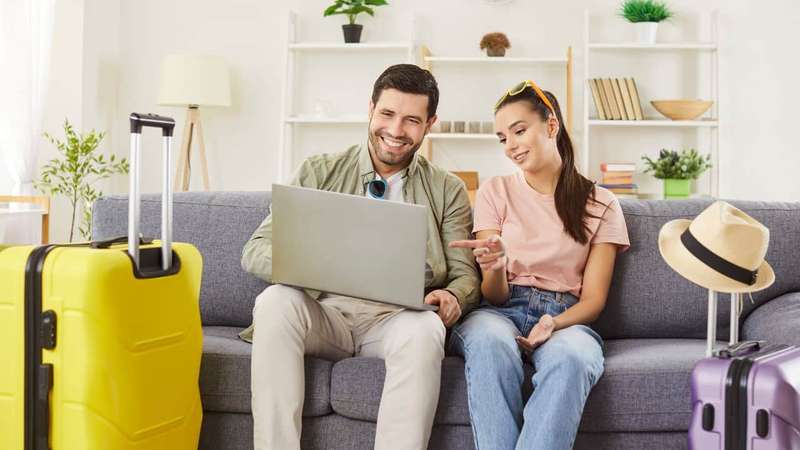 Newlywed couple planning their honeymoon trip, comparing prices on website while choosing tour online, enjoying summer holidays, posing on sofa with packed suitcase around. Travel concept