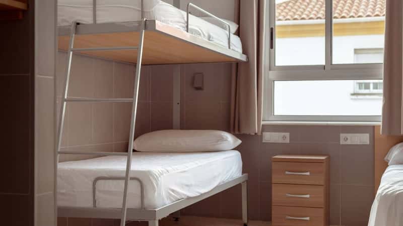 A clean and minimalistic hostel room with a bunk bed, white bedding, and a small wooden bedside table, ideal for budget travelers seeking affordable accommodation. Cheap hostels, save money.
