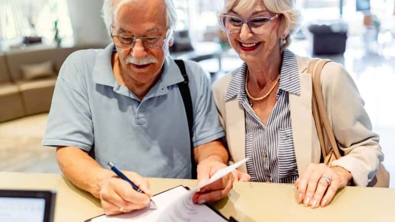 A smiling elderly couple stands in front of a hotel desk, filling out a check-in form or signing a bill, indicating they are either checking in or checking out after a wonderful stay.