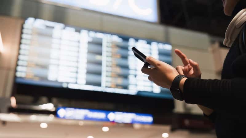 Woman in international airport, using mobile smartphone and checking flight schedule at the flight information board