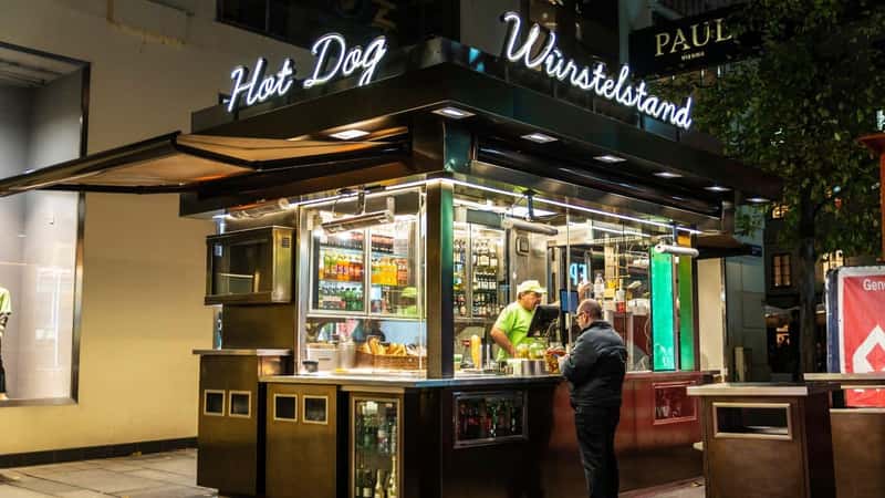 Vienna, Austria - October 16, 2022: Vendor in its sausage kiosk or Wurstelstand, food stand with people around at night in Innere Stadt, Vienna, Austria