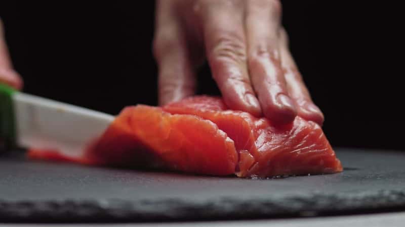 Sushi Chef Slices fresh Salmon on the sushi bar. Chef cutting salmon fillet at professional kitchen. Closeup chef hands slicing fresh fish slice in slow motion. Professional man cutting red fish