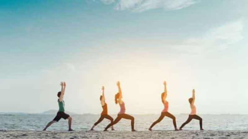 Young group man and woman yoga practice on mat relaxation in class exercise with pose fitness sport for healthy on the beach and seaside modern city at sunrise relax