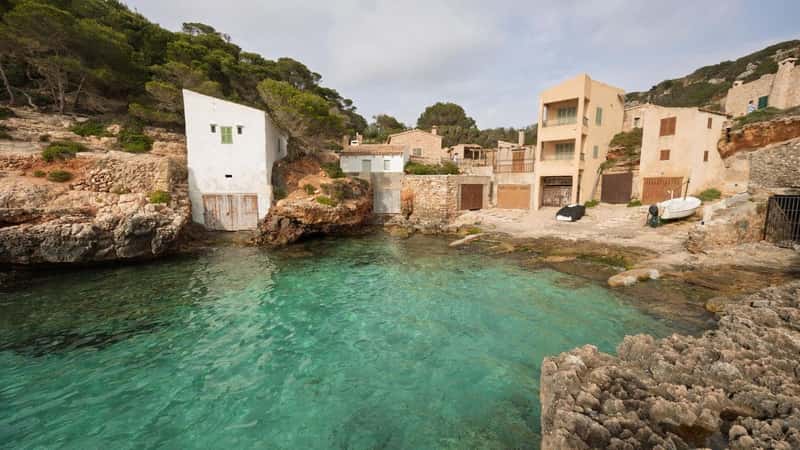 Crystal-clear turquoise water gently laps the shore of cala s'almunia, a charming cove in mallorca, where traditional houses nestle amidst the rocky landscape