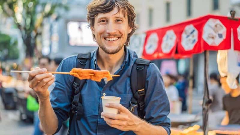 Young man tourist eating Typical Korean street food on a walking street of Seoul. Spicy fast food simply found at local Korean martket, Soul Korea