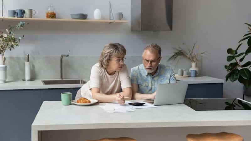 Serious older 60s married couple discussing budget, income, expenses, calculating money, counting insurance fees at calculator, doing paperwork together. paying bills, taxes, using laptop in kitchen