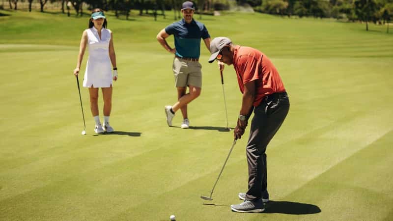 Golf players focus on putting as team members observe the competition on the green under a clear sky, highlighting teamwork and precision.