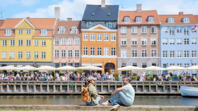 Two people sit on a cobblestone walkway along a canal in Copenhagen, enjoying the sunshine and a view of colorful buildings. Copenhagen Denmark