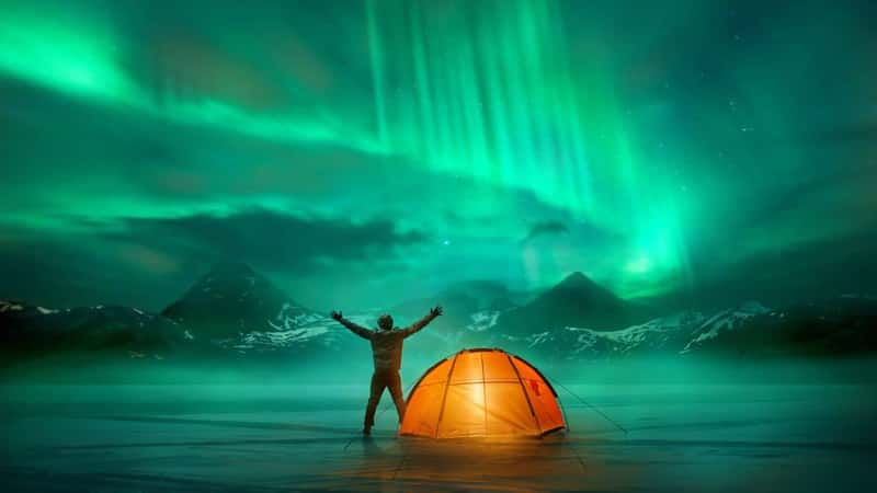 A man camping in wild northern mountains with an illuminated tent viewing a spectacular green northern lights aurora display. Photo composition.