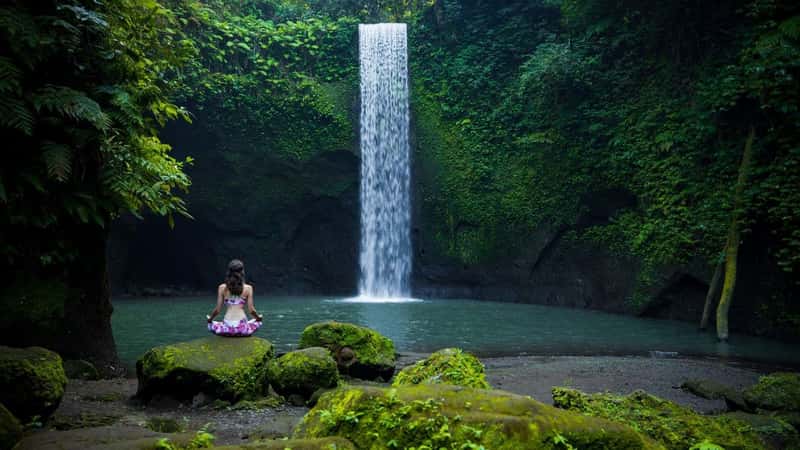 Yoga lotus pose. Young Caucasian woman sitting on the stone, meditating, practicing yoga, pranayama at waterfall. Hands in gyan mudra. Yoga retreat. View from back. Tibumana waterfall, Bali, Indonesia