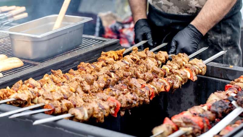 Chef in black gloves cooking meat kebabs on grill. Marinated meat skewers with vegetables street food preparation at barbecue festival.