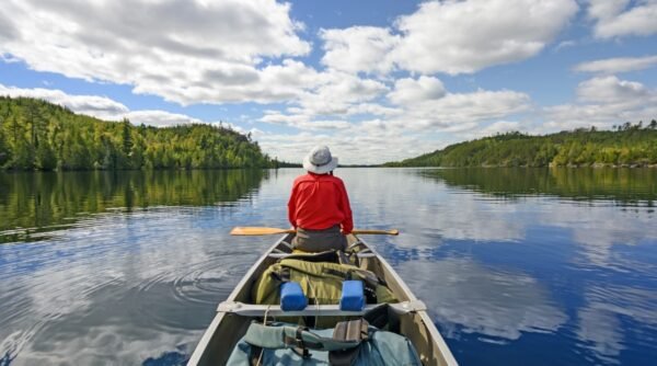The view to the canoeist on Kekekabic Lake in the Boundary Waters of Minnesota. The pristine waters of the lake, surrounded by dense forests and framed by the rugged silhouette of distant hills. The mirror-like surface of the lake reflects the lush greenery and the blue sky with clouds. In a wooden canoe lies a lot of different things and sits a canoeist dressed in gray trousers, a red shirt and a light gray Panama hat.