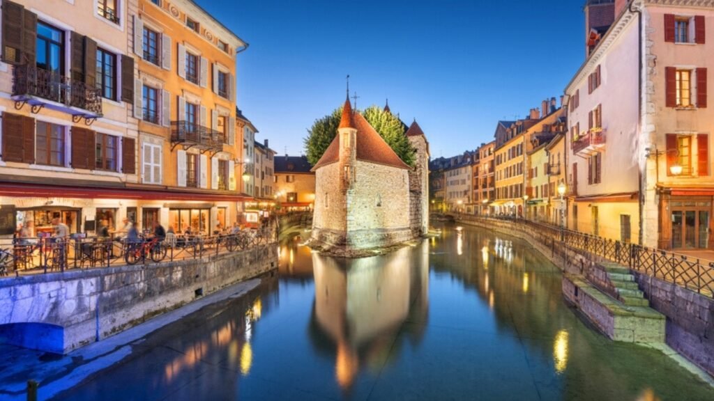 The Palais de I'lle is a picturesque medieval fortress located on an island in the Thiou River, in the heart of Annecy, France. This historic building boasts a unique and charming appearance, featuring a distinctive triangular shape with turrets and a sloping, tiled roof. he surrounding environment is equally enchanting, with cobblestone streets, quaint cafes, and the pristine waters of the Thiou River.